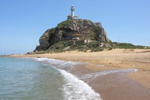 Paisaje costero con un faro en un promontorio rocoso, playa y océano.