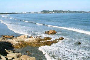 Una costa rocosa con olas, edificios lejanos y un cielo despejado.