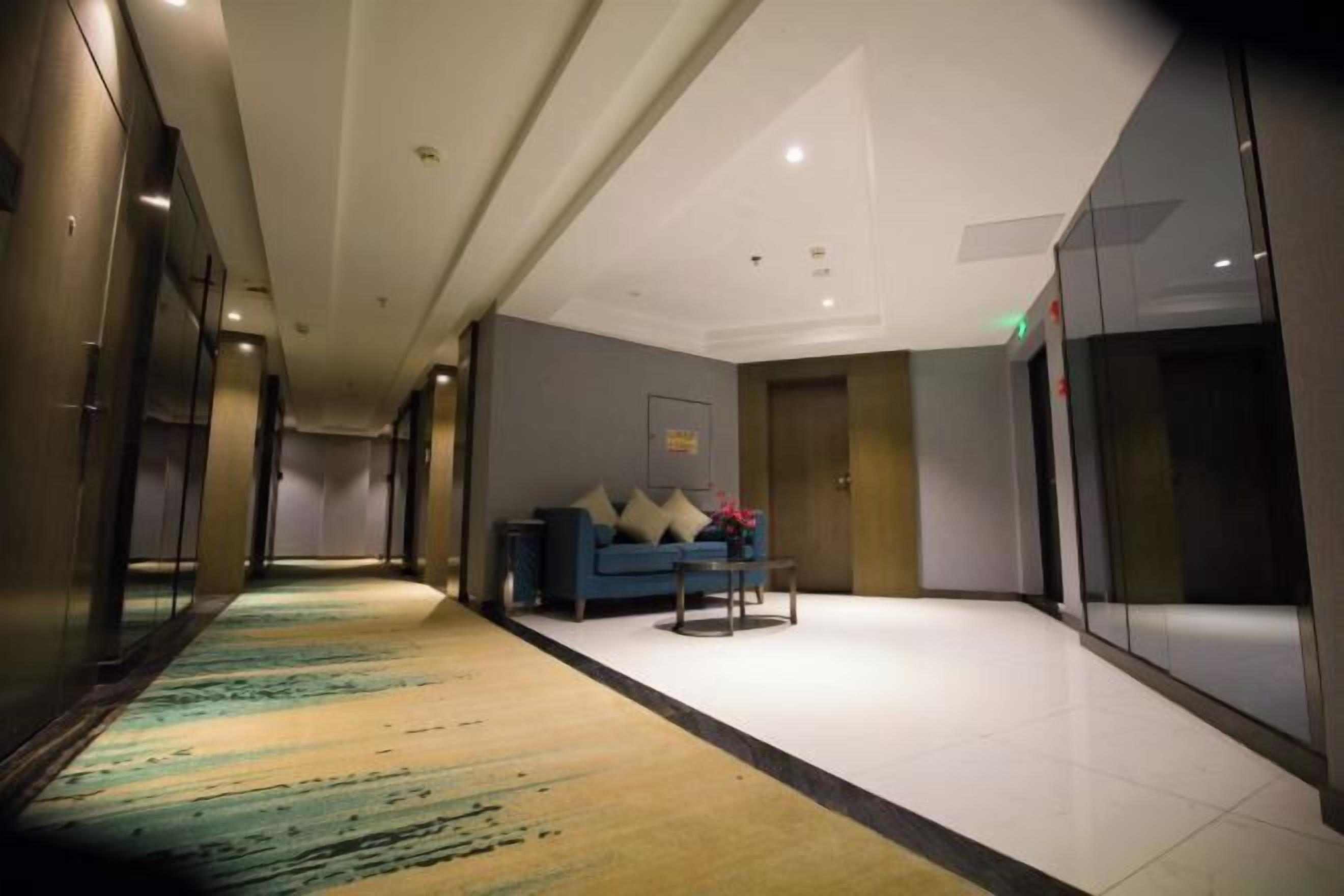 Un pasillo de hotel con alfombra estampada, un sofá azul con cojines y una pequeña mesa.
