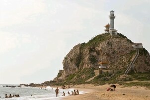 Una playa con un faro en un acantilado rocoso, gente tomando el sol y un edificio con techo inclinado.