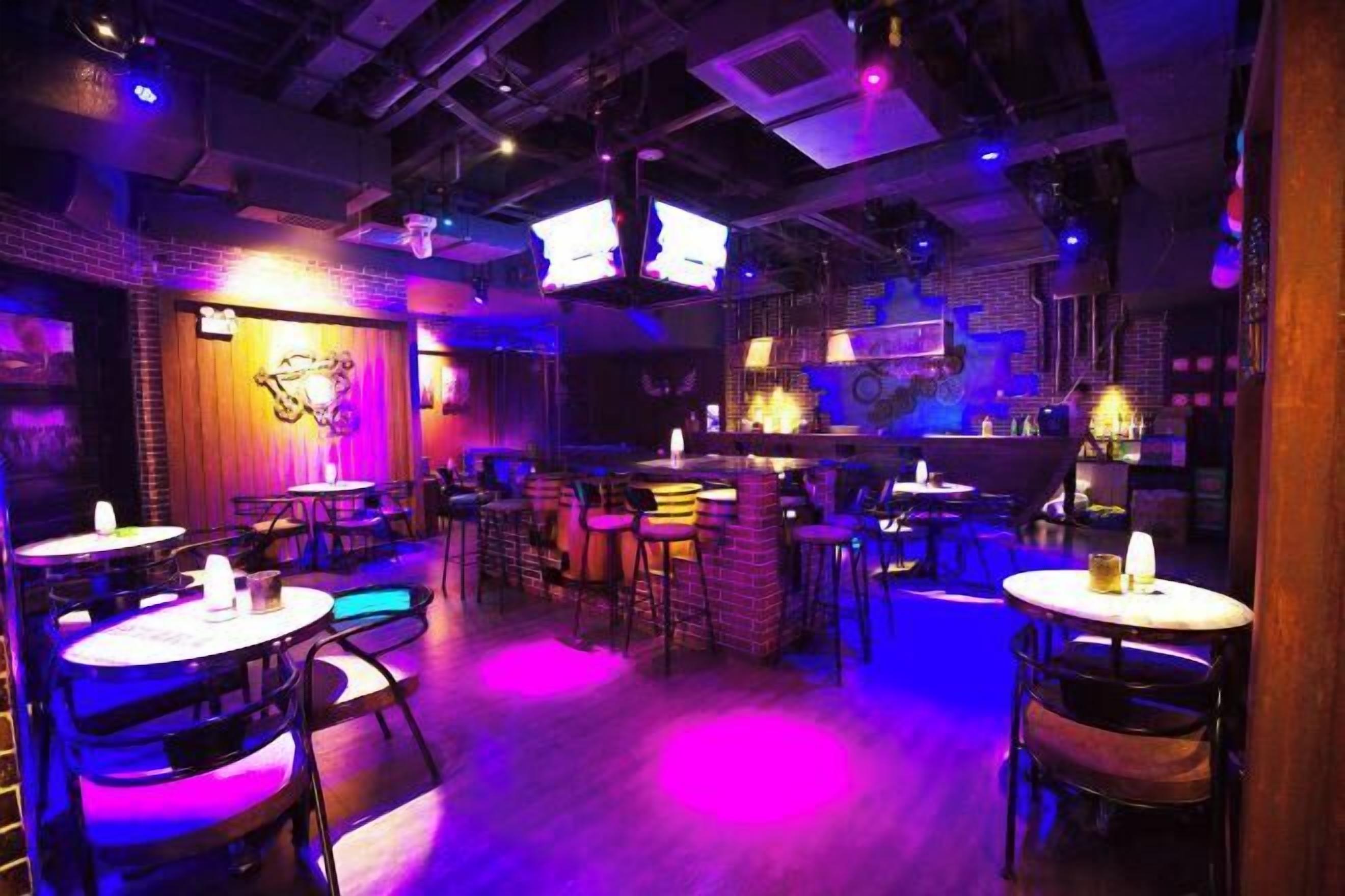 Eine Bar mit mehreren Tischen und Hockern, einer Ziegelwand mit einem Neonzeichen und einer Bühne mit DJ-Equipment.