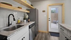 Cocina compacta con pileta, lavavajillas y refrigerador. Hay repisas de madera sobre la encimera y una puerta que da a un patio.