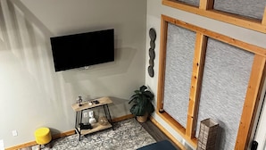 Una sala de estar con un sofá azul oscuro, un televisor de pantalla plana montado en la pared, una pequeña mesita de madera y una planta en maceta.