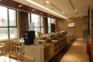 Zona de desayuno de hotel con una máquina de café, bufé, asientos y una pantalla de televisión.