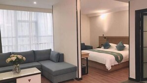 A modern hotel room with a grey sofa, a small table with a vase of flowers, a bed with green bedding, and a wooden floor.