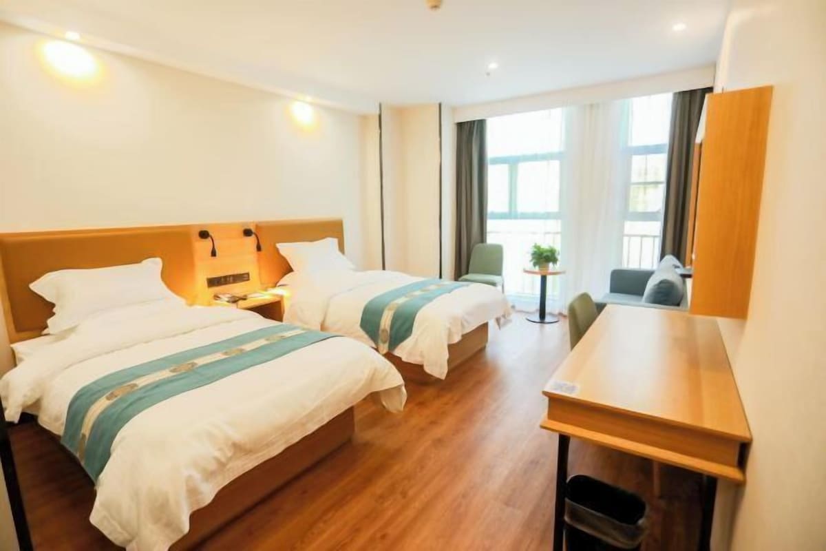 Quarto de hotel com duas camas, uma escrivaninha, uma cadeira, uma mesinha e uma janela com cortinas.
