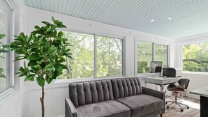 A modern office space with a grey sofa, a desk with a computer, and a large window with a view of trees.