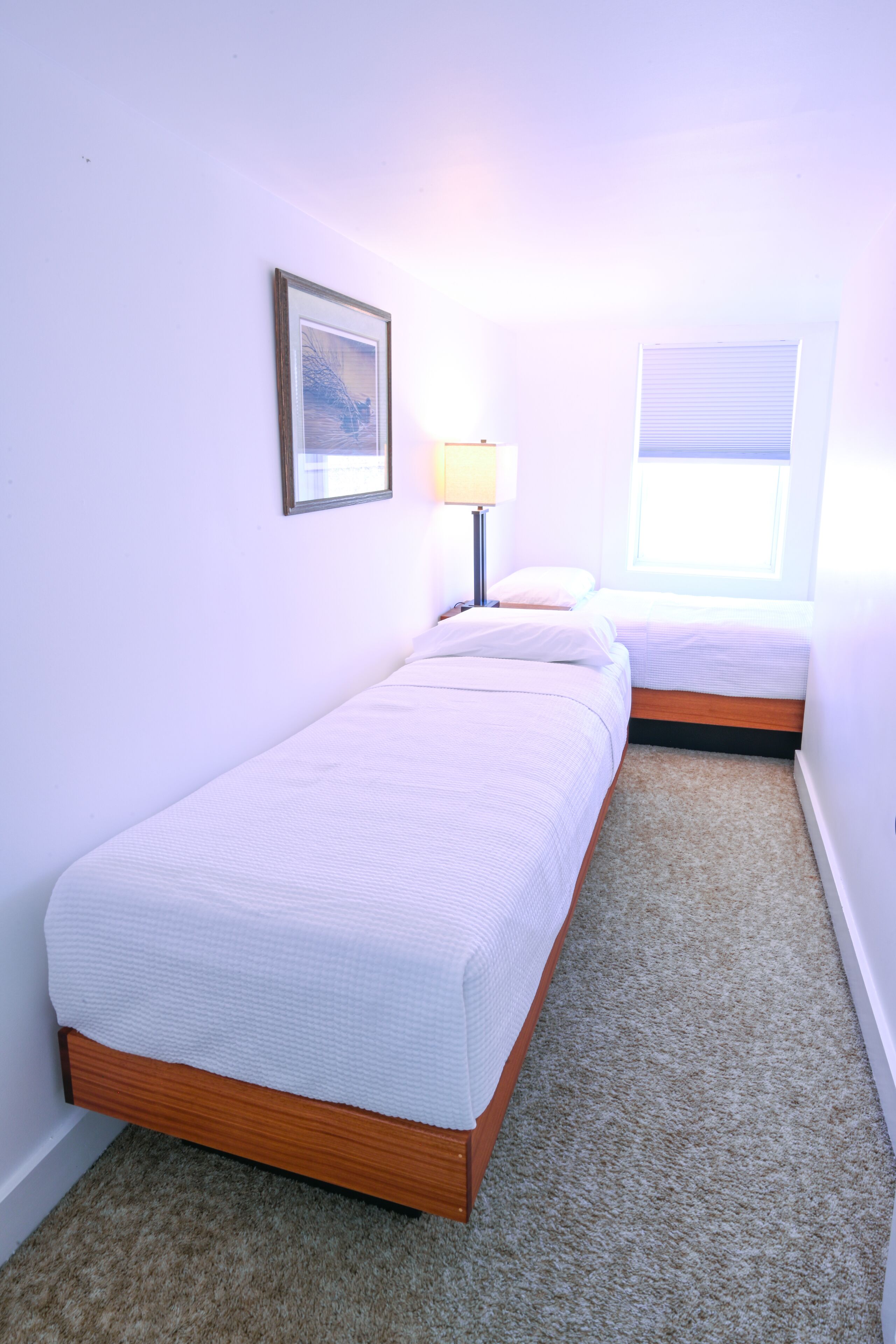 A bedroom with two single beds, a framed picture on the wall, a lamp, and a window with blinds.