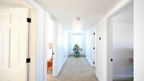 Un pasillo con paredes blancas y alfombra que conduce a una habitación con ventana y planta.