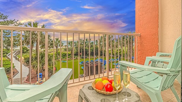 Un balcón con vista a una piscina y palmeras, dos sillas verdes y un frutero con bebidas.