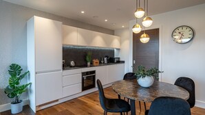 Een moderne keuken met witte kasten, een donker aanrecht en een houten eettafel met zwarte stoelen. Boven de tafel hangen hanglampen en er is een klok aan de muur.