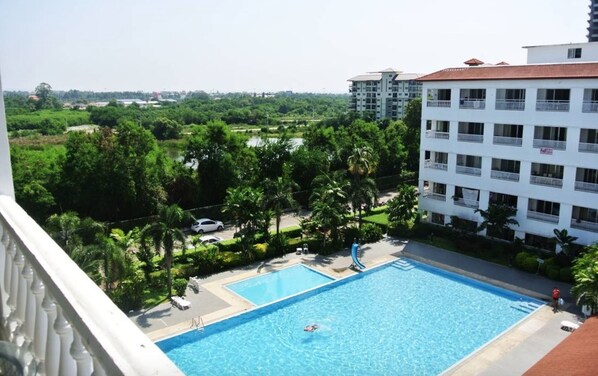 A swimming pool with a person swimming, surrounded by greenery and a white building with balconies.
