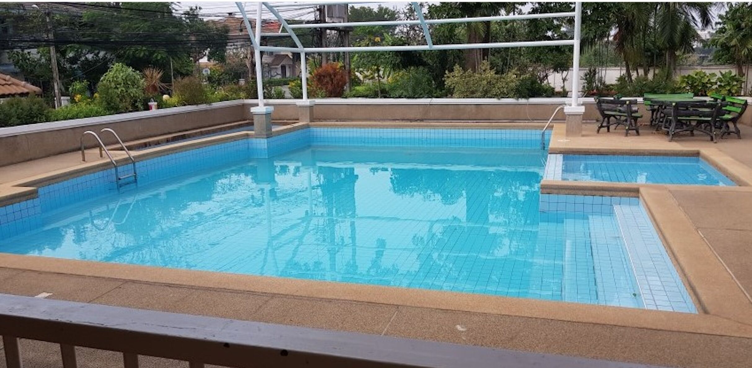 A rectangular swimming pool with a tiled edge, a metal ladder, and a small step leading into the pool. There are outdoor tables and chairs to the right.