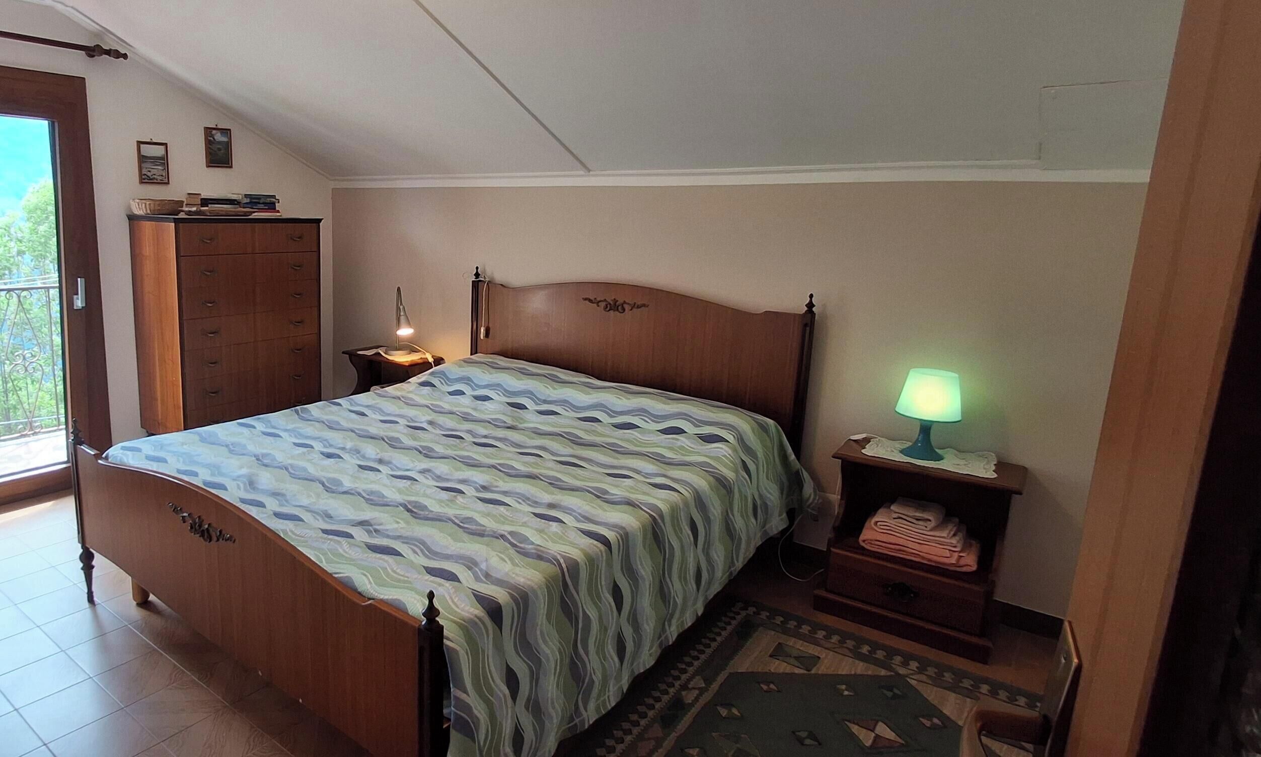 Ein Schlafzimmer mit einem hölzernen Bett, einem Nachttisch mit Lampe, einer Kommode und Blick ins Freie.