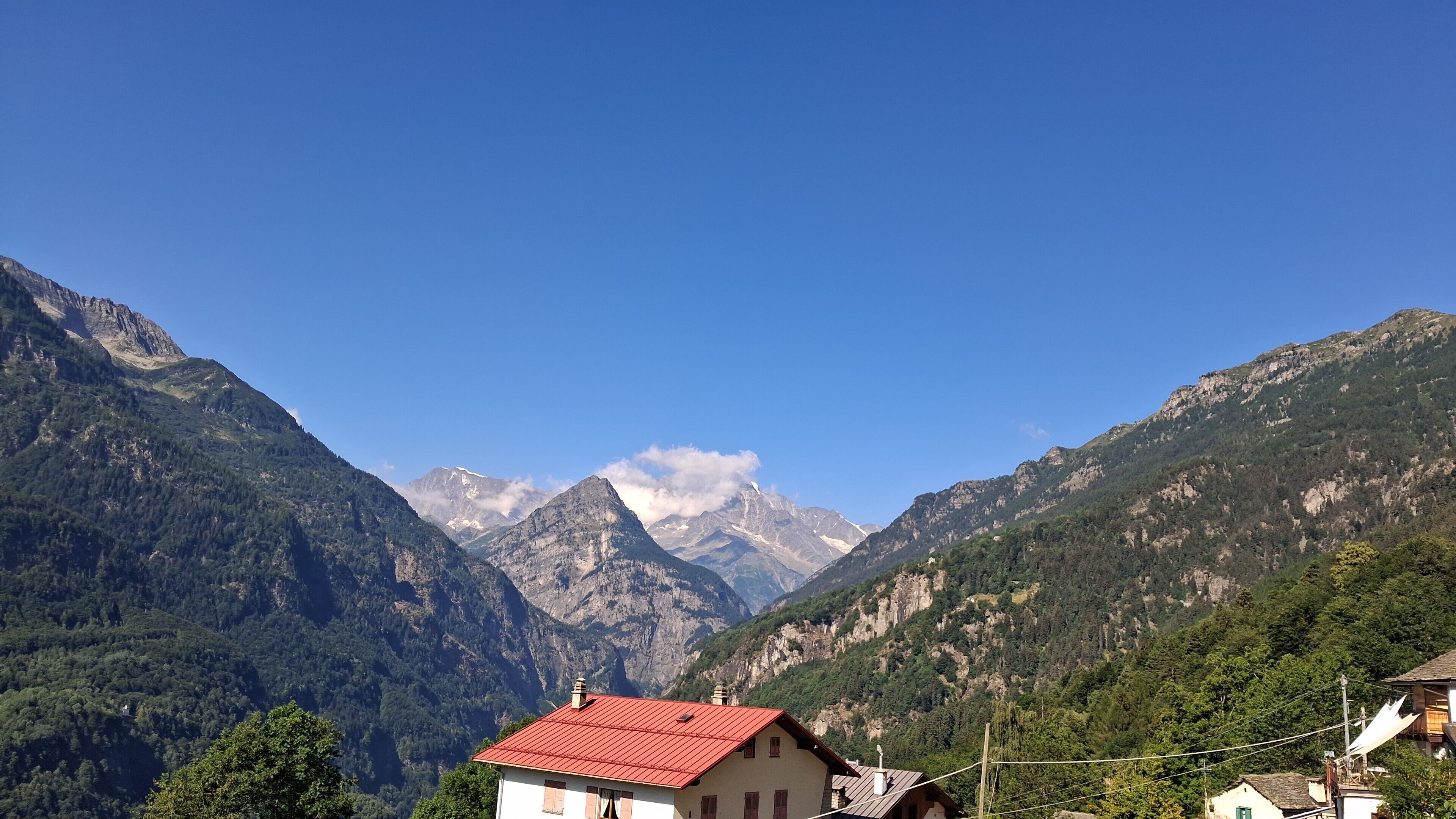 A mountain landscape with a house featuring a red roof, surrounded by lush greenery and mountains.