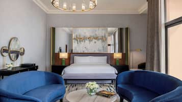 Habitación de hotel con una cama grande, dos sillones azules, una mesa redonda de centro con un jarrón y un libro, una araña y un espejo grande.