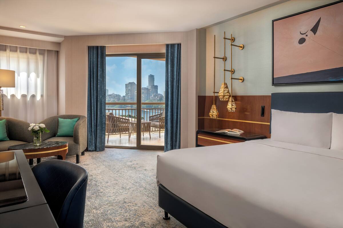 Habitación de hotel con una cama grande, un escritorio con silla, un sofá con almohadas, vistas de la ciudad desde un balcón y un cuadro en la pared.