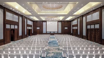 Uma sala de conferências grande com fileiras de cadeiras brancas, carpete azul, painéis de madeira e uma tela grande na frente.