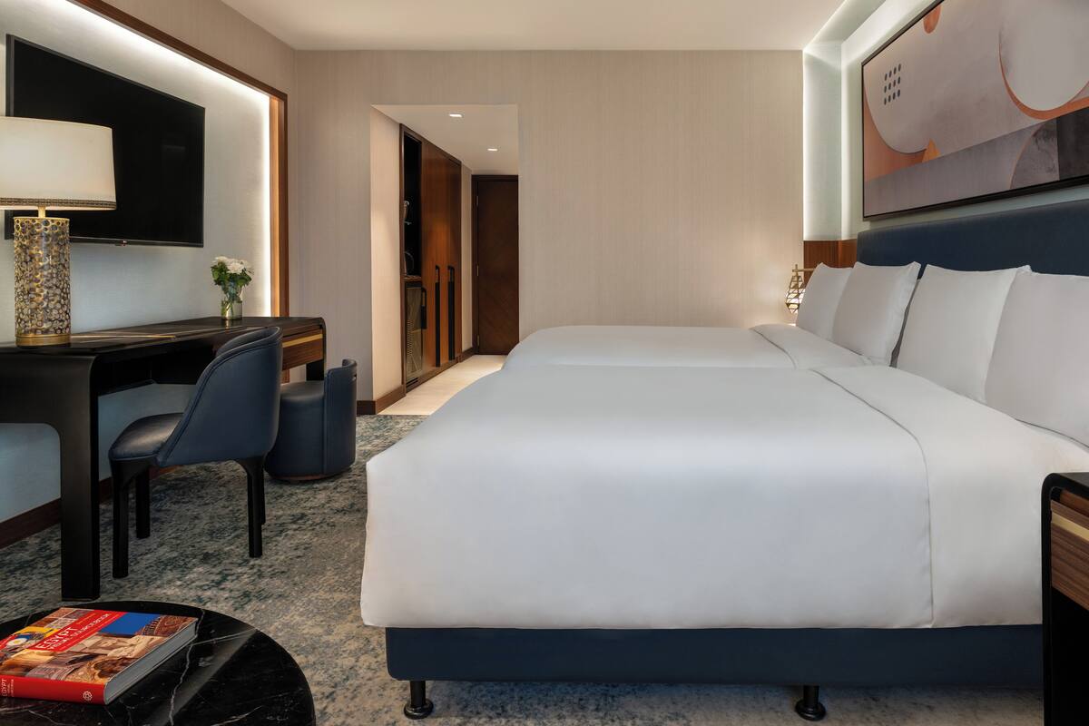 Una habitación de hotel con una cama grande, un escritorio con silla, un televisor montado en la pared y una mesita de noche con lámpara y un libro.