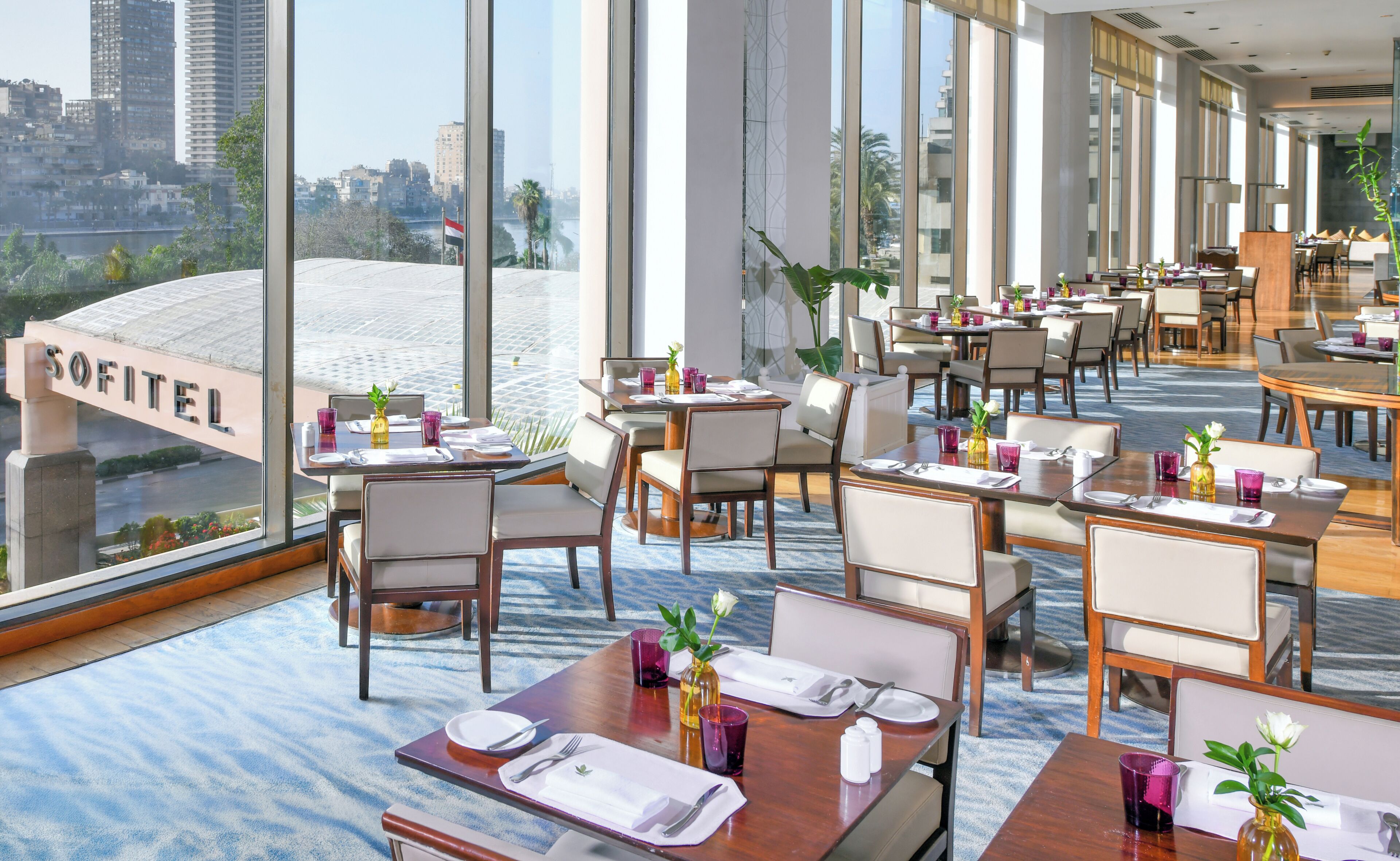 A modern restaurant with tables set for dining, large windows offering a city view, and a sign reading 'SOFITEL'.