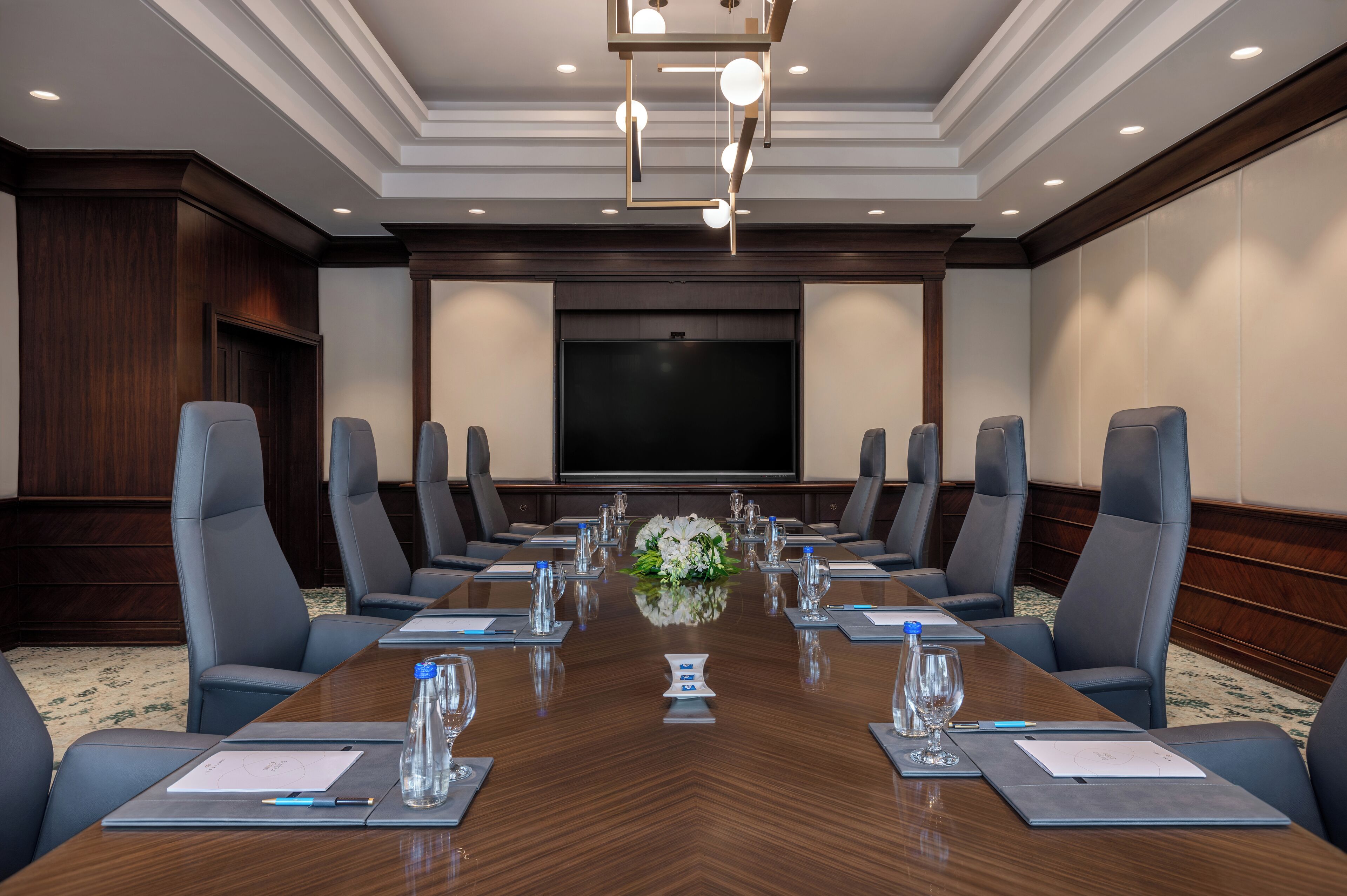 A conference room with a long table, multiple chairs, a large screen, and a floral centerpiece.
