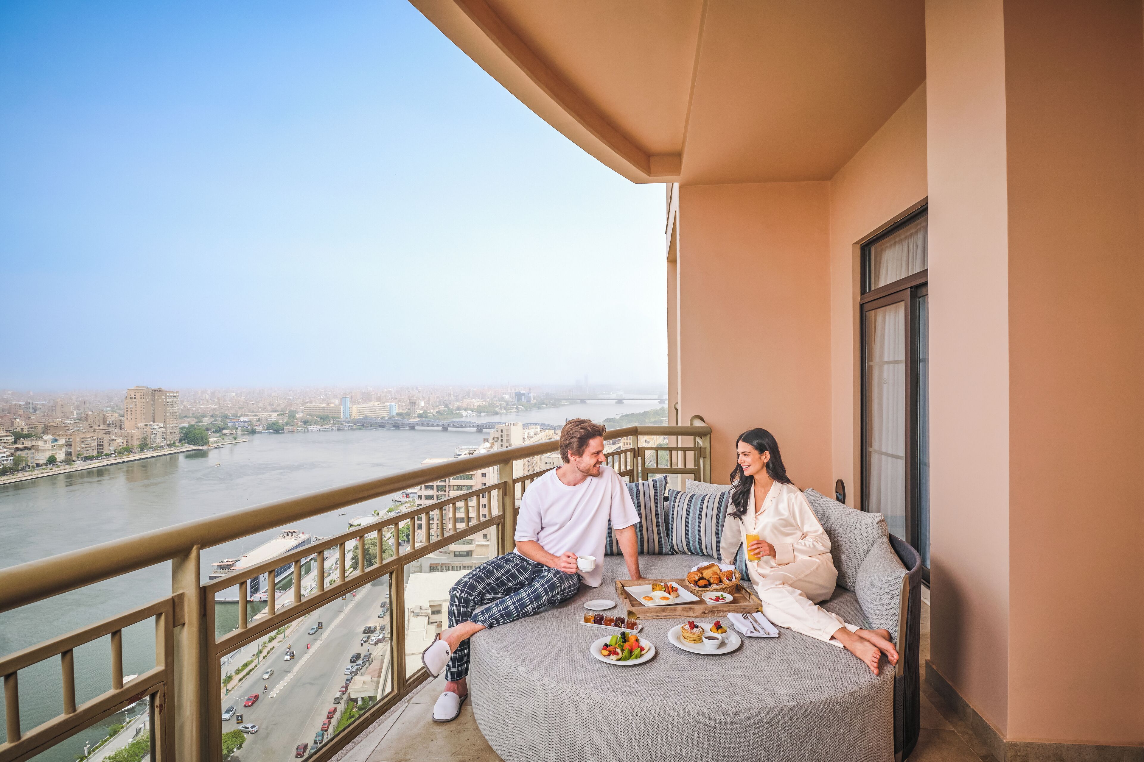 A couple on a balcony with a city view, dining table with food, and a railing.