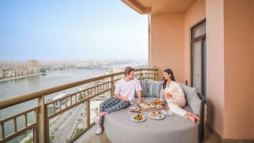 A couple on a balcony with a city view, dining table with food, and a railing.