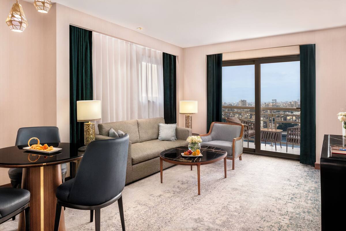 Una habitación de hotel moderna con un sofá, dos sillones, una mesa de centro y una mesa de comedor. Hay dos lámparas, un jarrón con flores y vistas a la ciudad a través de amplios ventanales.
