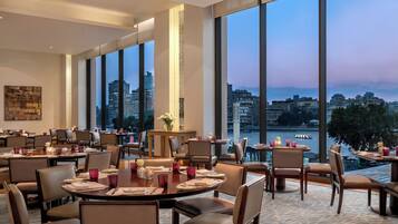 A restaurant with large windows offering a city view, multiple dining tables, and chairs.