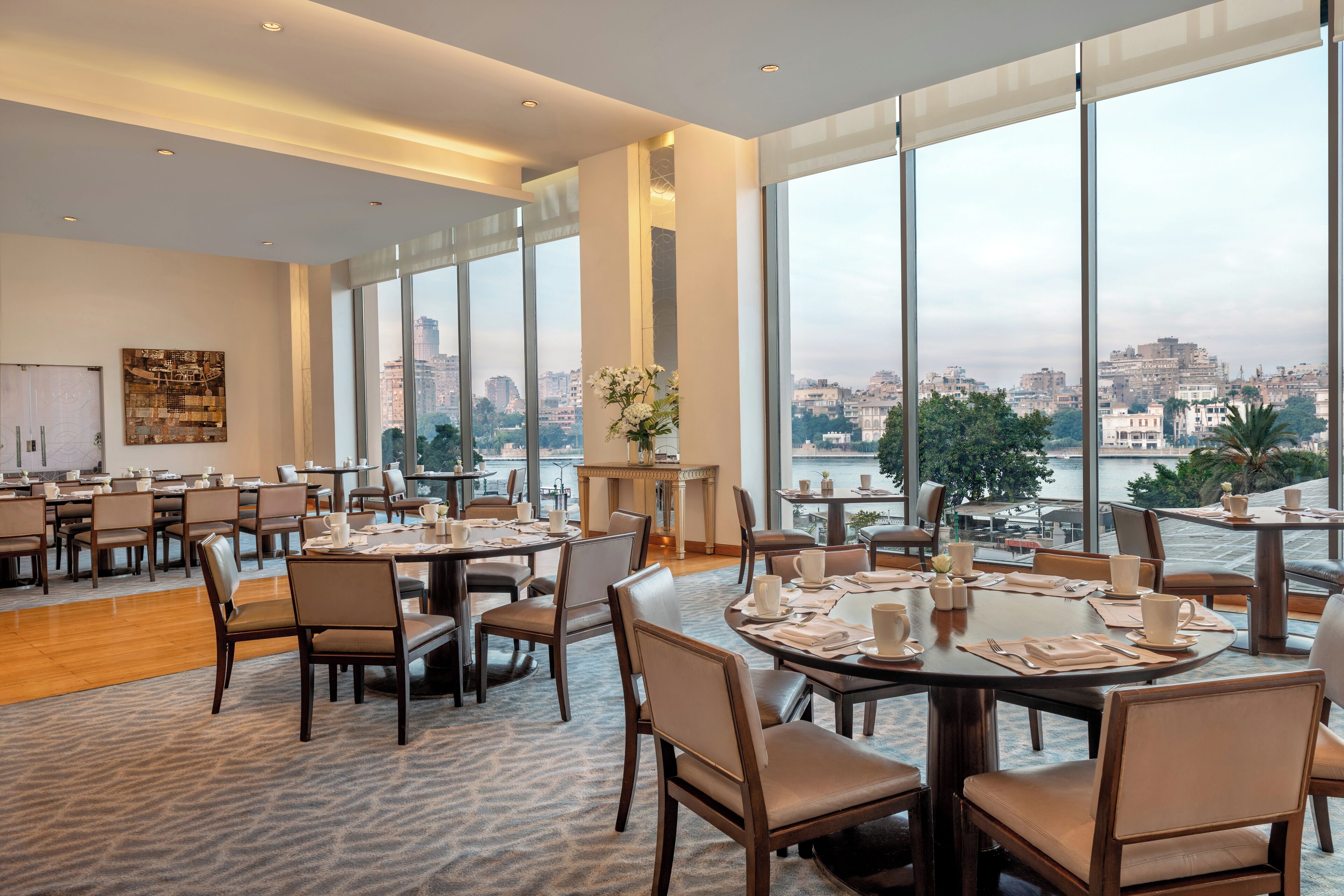 Un vaste espace dédié à la salle à manger, doté de grandes fenêtres offrant une vue sur la ville. Les tables sont disposées avec soin et accompagnées de chaises, et la pièce présente un design moderne avec un sol recouvert de moquette.