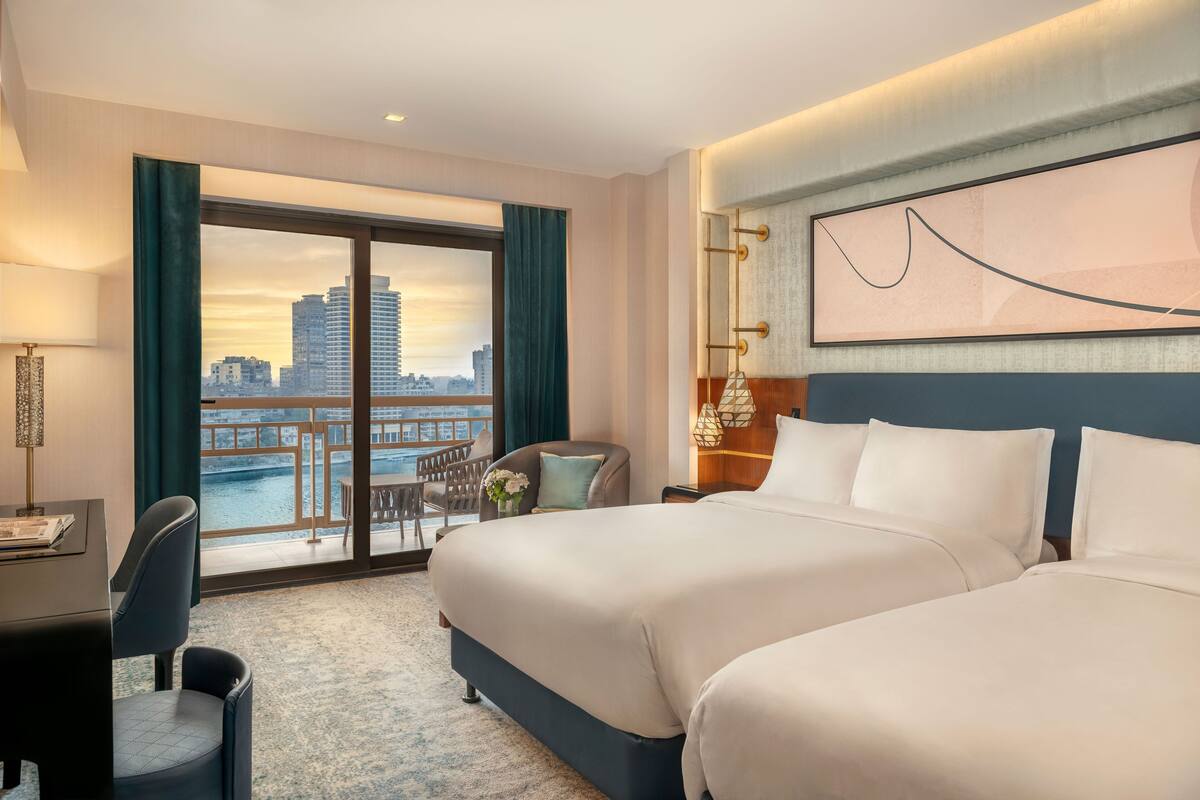 Habitación de hotel con dos camas, un escritorio, una silla, vista a un paisaje urbano a través de un ventanal grande y un cuadro abstracto enmarcado en la pared.