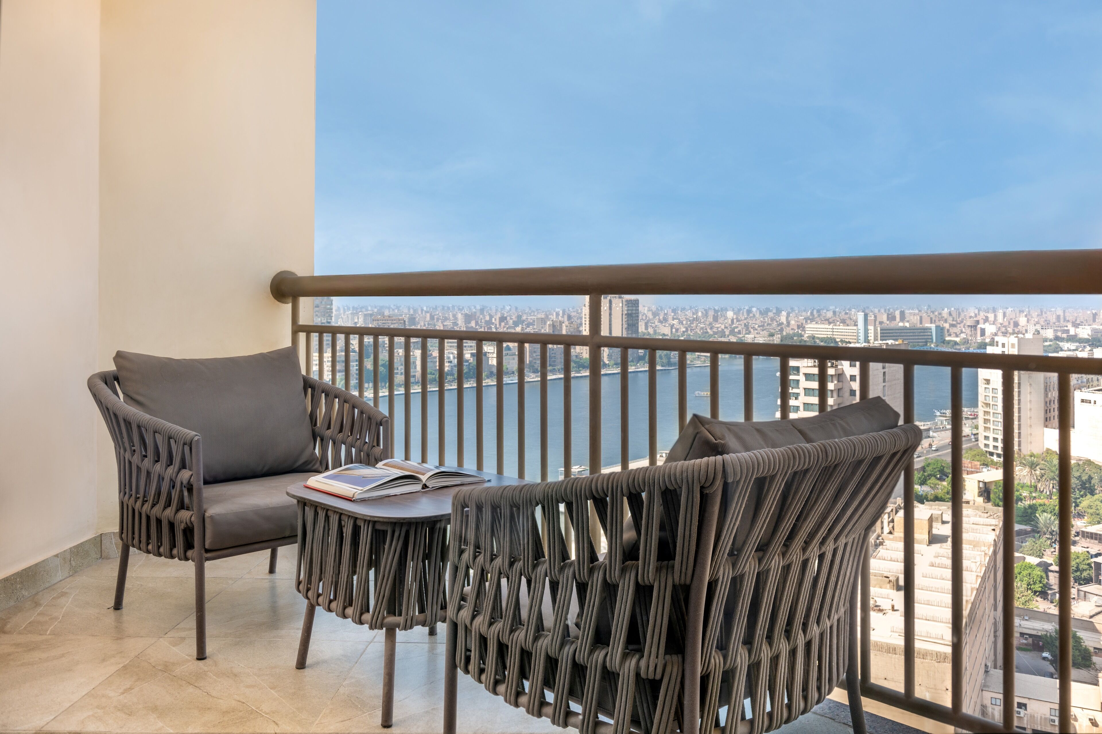 A balcony with two chairs and a small table, overlooking a cityscape and a body of water.