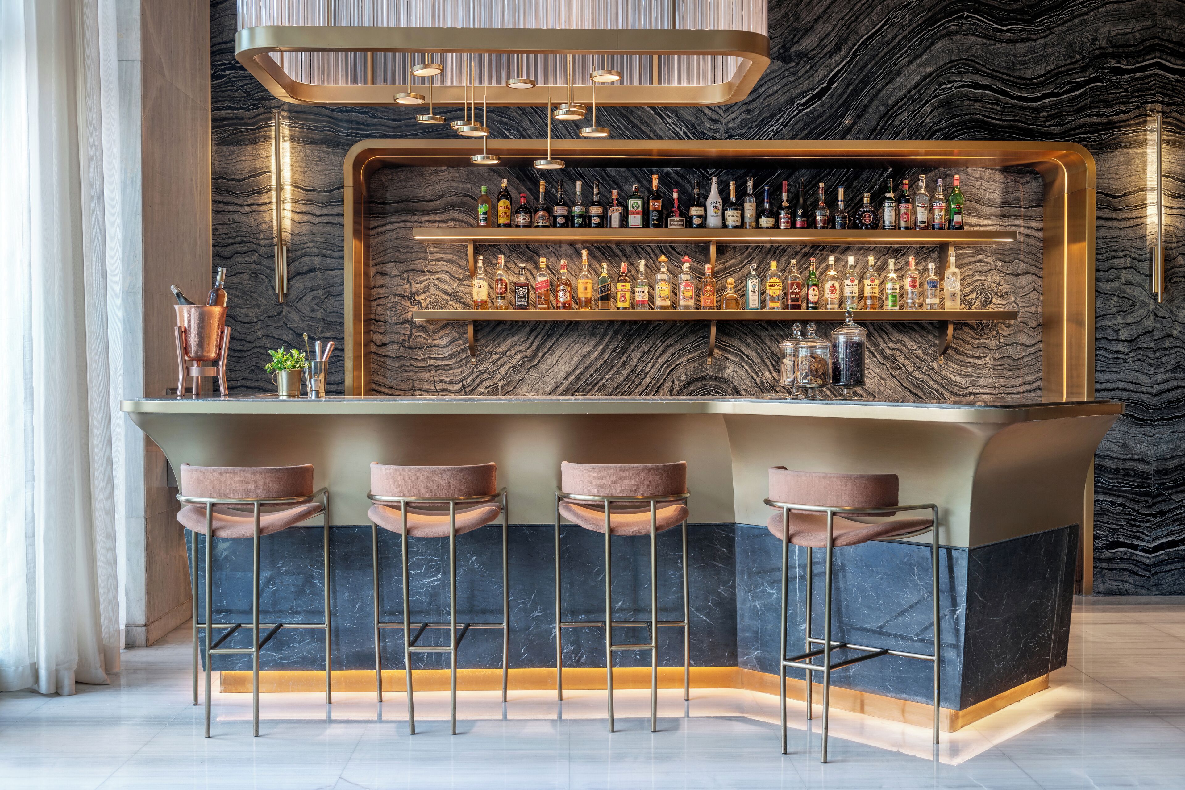A bar area with a marble wall, multiple shelves of liquor bottles, and three bar stools.
