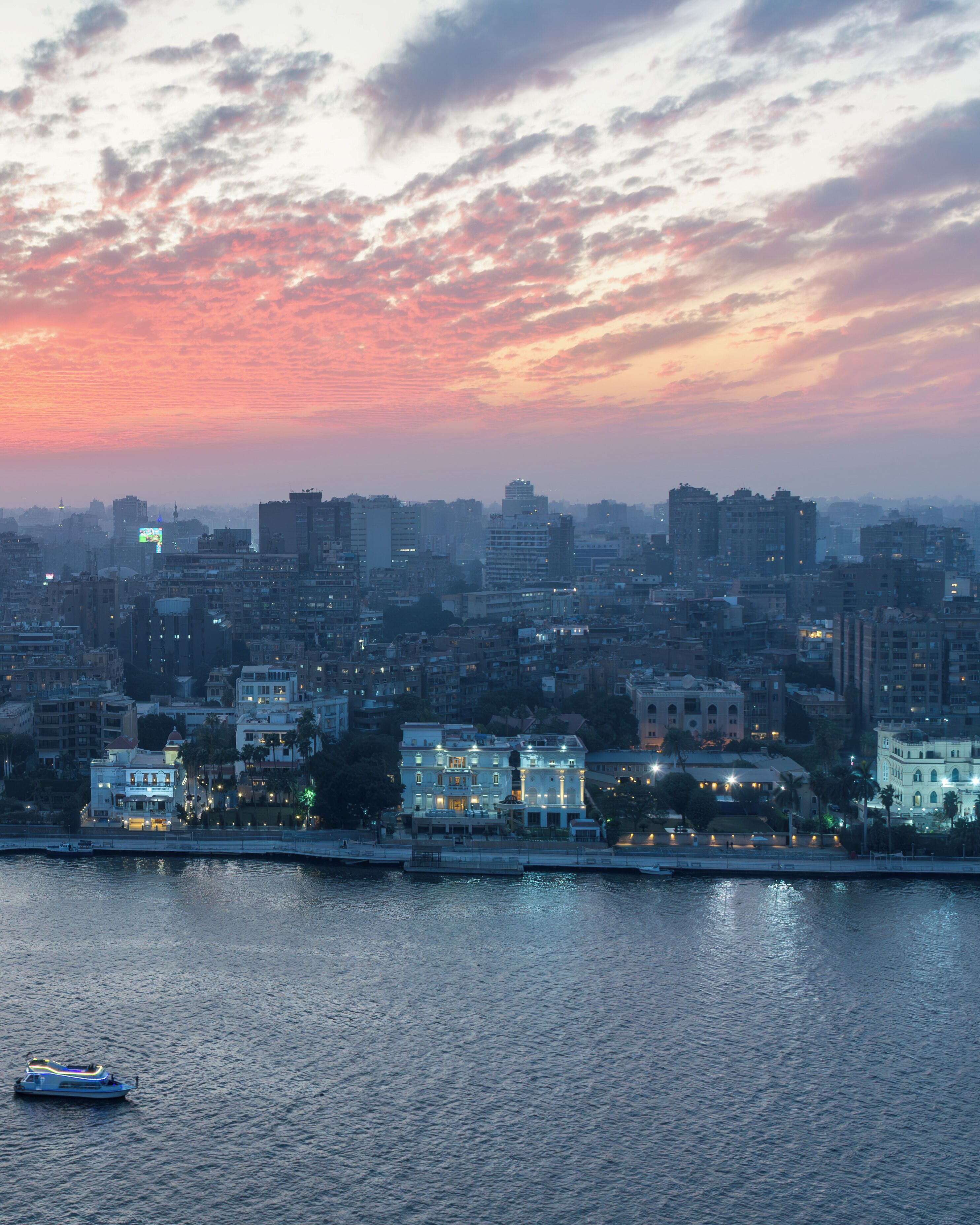 A cityscape at dusk with buildings along a waterfront, a boat on the water, and a bridge in the foreground.