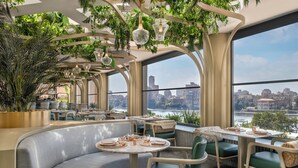 A restaurant with a view of a city skyline and a river. It features round tables, green cushioned seating, and hanging plants.