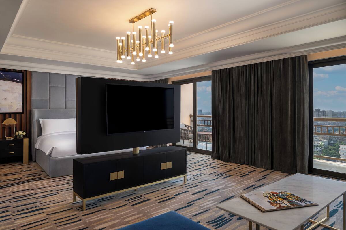 Una habitación de hotel moderna con una cama grande, un televisor de pantalla plana, una mesa de comedor y vistas a la ciudad a través de amplias ventanas.