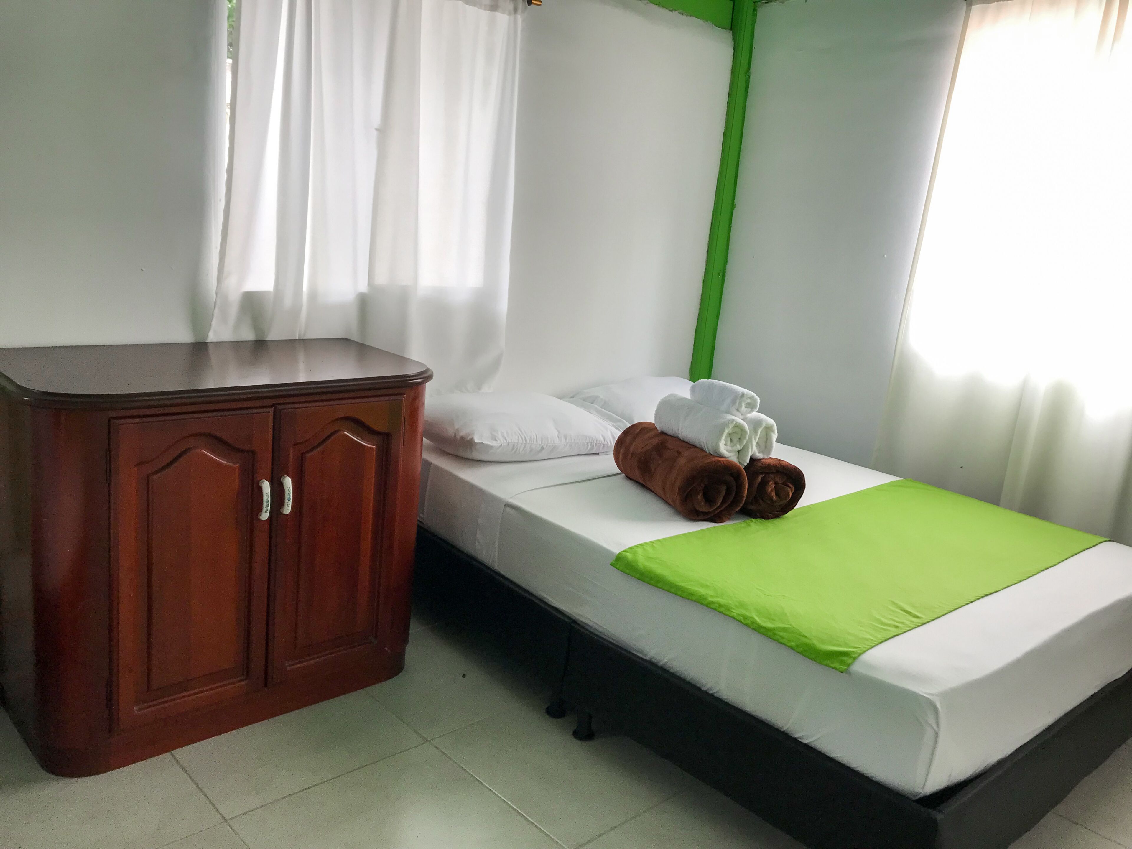 A single bed with a green bedspread, white pillows, and rolled towels.