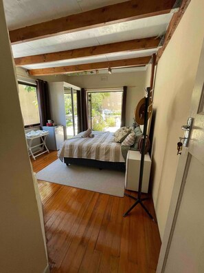 A bedroom with a wooden floor, a bed with a white and gray striped bedspread, a bedside table with a lamp, and a view of a garden through a large window. - Room-living In Mountain Views Camps Bay (Cape Town)