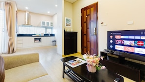Uma sala de estar moderna com um sofá, uma mesa de centro com uma revista e flores, uma TV exibindo serviços de streaming e uma cozinha ao fundo.