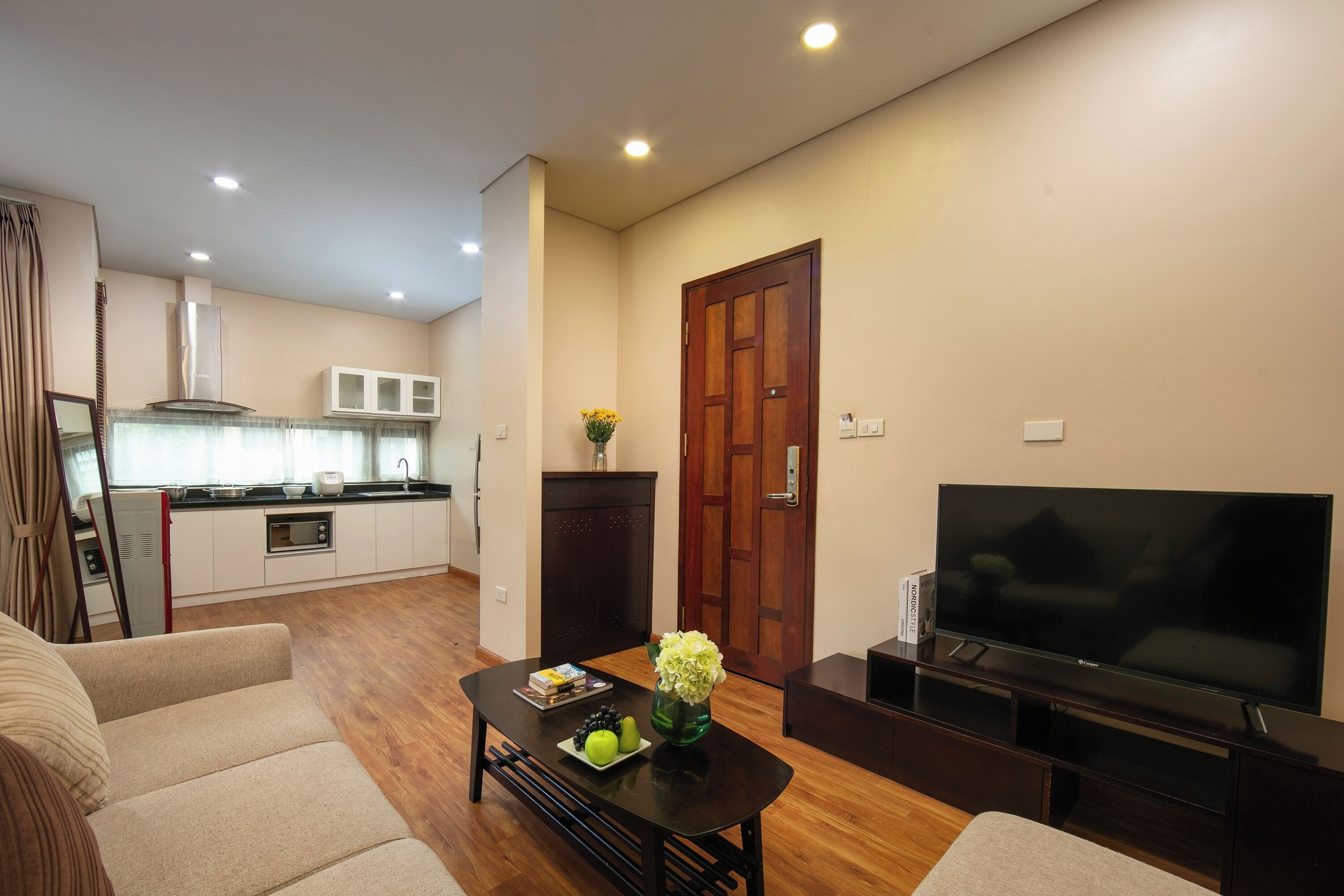 Una sala de estar moderna con un televisor de pantalla plana, una mesa de comedor con libros y fruta, y una cocina al fondo.