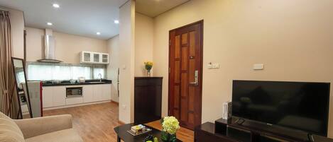 Una sala de estar moderna con un televisor de pantalla plana, una mesa de comedor con libros y fruta, y una cocina al fondo.