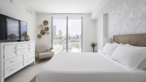 A modern bedroom with a large bed, a TV, a white dresser, and a city view through the window. - Artistic Design District Stay, Parking, Pool, Gym (Miami)