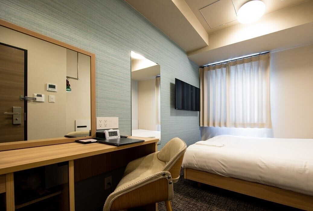 Una habitación de hotel con una cama, un escritorio con una silla, un televisor montado en la pared y una ventana con cortinas.