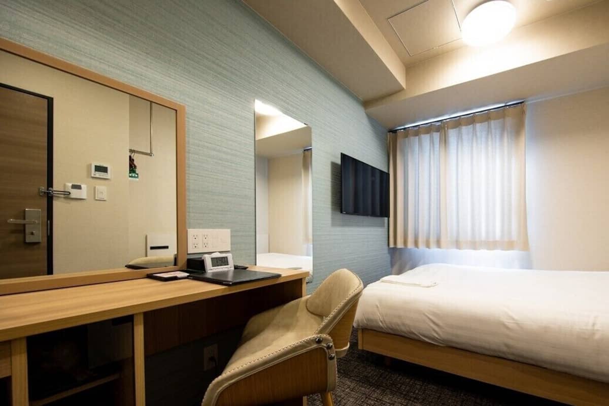 A hotel room with a bed, a desk with a chair, a TV mounted on the wall, and a window with curtains.