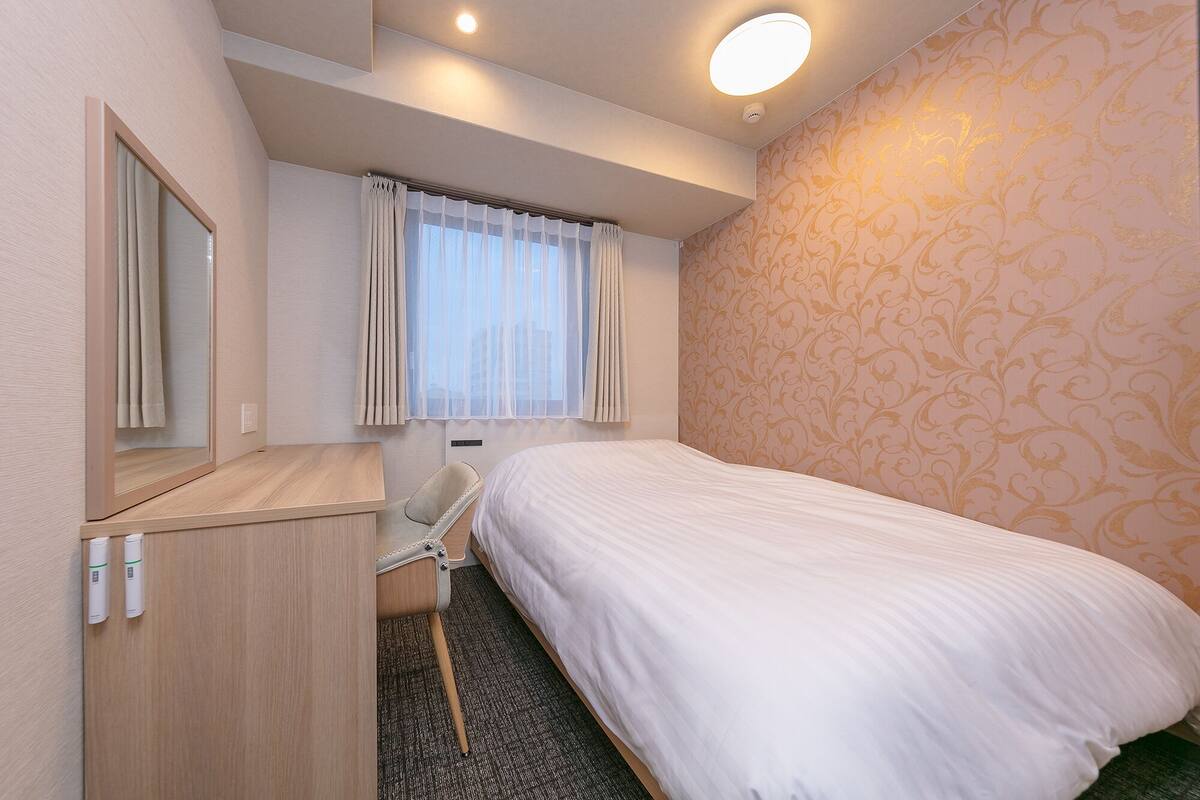 A hotel room with a bed, a desk, a chair, a mirror, a window with curtains, and a patterned wall.