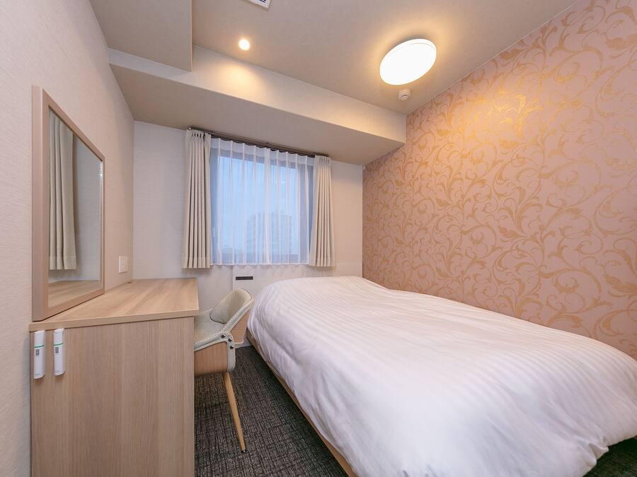 A hotel room with a bed, a desk, a chair, a mirror, a window with curtains, and a patterned wall.