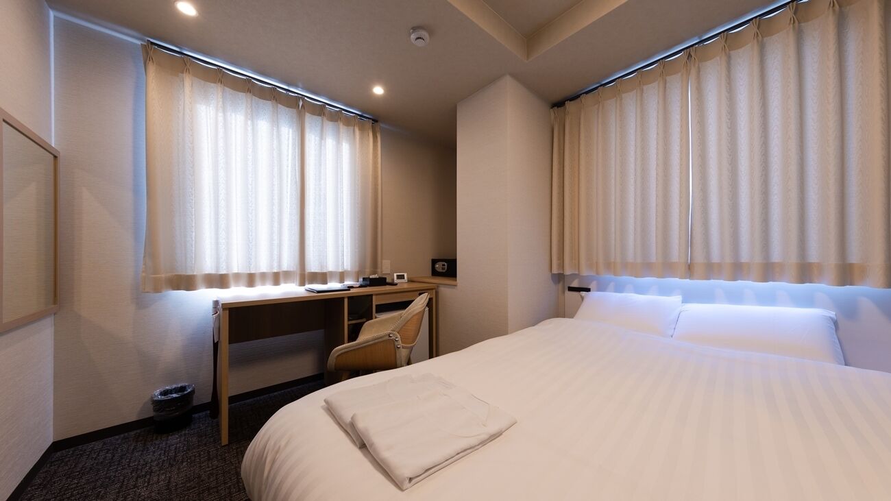 Habitación de hotel con cama, escritorio, silla y dos ventanas con cortinas.