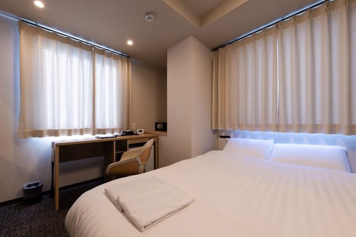 A hotel room with a bed, a desk, a chair, and two windows with curtains.