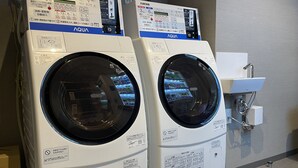 Two front-loading washing machines with control panels, placed in a room with a sink and a tiled floor. - Remington Hotel (Akashi)
