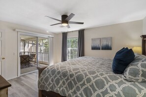 A bedroom with a large bed, a ceiling fan, a balcony with outdoor furniture, and a view of the outdoors.